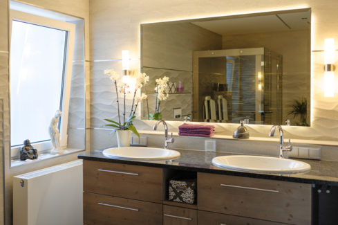 Badezimmer mit Natursteinwänden, einem großen Spiegel in der Mitte von 2 Waschbecken, eine Orchidee ist links neben dem linken Waschbecken